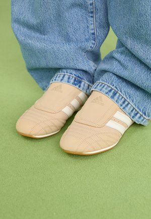 Beige sportskor med texturerad ovandel och kardborreband, med tre vita ränder och gummisula, i kombination med blåa denimjeans.