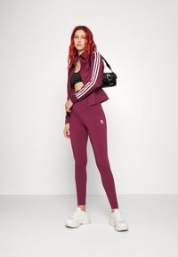 Vínová sportovní souprava s třemi bílými pruhy na rukávech bundy. Černý crop top pod tím a bílé tenisky. Modelka drží černou tašku.