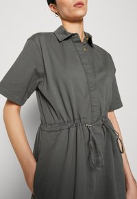 Personne portant une robe grise à manches courtes avec col et taille ajustable à cordon, boutons dorés et boucle d'oreille en anneau doré, devant un fond uni.