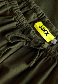 Shorts vert olive avec une taille élastique, un cordon de serrage et une étiquette de marque jaune. Le tissu semble léger avec une texture lisse.