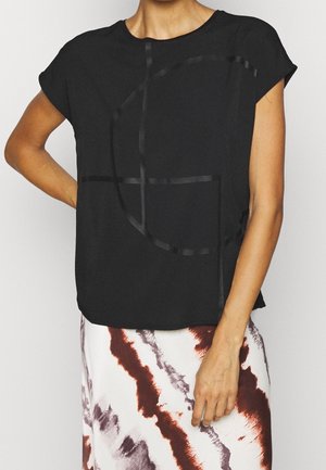 Femme portant un haut à manches courtes noir avec un motif géométrique brillant et une jupe blanche avec des stries abstraites marron et violettes, bras plié à la taille.