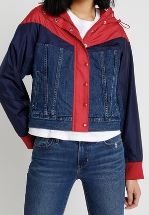 Woman wearing a cropped jacket combining red and navy nylon with blue denim panels, paired with blue jeans and a white shirt underneath.