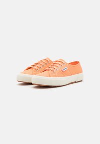 Orange canvas sneakers with white rubber sole, round toe, and metal eyelets. Features a textured upper and small logo patch on the side.