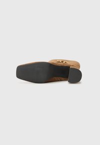 Sapato de salto em camurça castanha com sola de borracha preta lisa; possui um detalhe em metal dourado circular na parte superior do sapato.