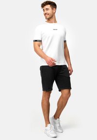 Camiseta blanca con logotipos negros en las mangas combinada con pantalones cortos de color gris oscuro. El modelo lleva zapatillas blancas. Ajuste casual y cómodo.