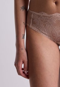 Mutande hipster beige in pizzo con motivi floreali, tessuto testurizzato ed estremità delicate, viste accanto a una mano parzialmente visibile.