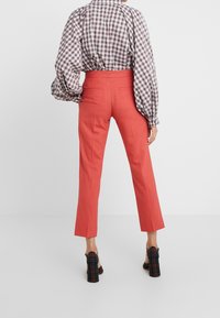 Žena v korálových upravených zkrácených kalhotách, kostkované dlouhé košili s nabušenými rukávy a kostkovaných vysokých podpatcích, stojí against bílém pozadí.