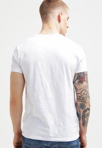 Hvid bomulds t-shirt, korte ærmer, rund hals. Glat tekstur, afslappet pasform. Synlige farverige tatoveringer på armen.