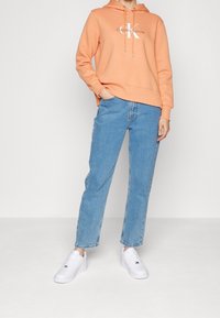 Light orange hooded sweatshirt with logo, paired with blue straight-leg jeans and white sneakers, featuring a simple design and relaxed fit.