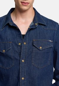 Jeanshemd in Dunkelblau mit zwei Brusttaschen, goldenen Knöpfen und einem spitzen Kragen. Verfügt über einen strukturierten Stoff und sichtbare Nähte.