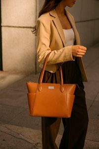 Vrouw in een beige blazer en witte top, met een bruin leren handtas, die op de stoep van de stad loopt in warm licht.