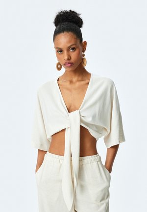 Witte cropped top met een fronttie, korte mouwen en een textuurstof. Model draagt beige broeken met een elastische tailleband.