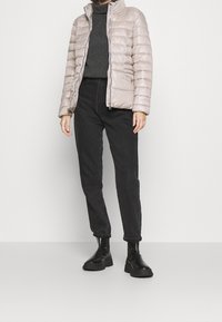 Wypełniona kurtka w jasnym odcieniu beżowym z wysokim kołnierzem, zamek błyskawiczny oraz poziomy wzór pikowania, noszona na szarym golfie oraz czarnych spodniach o prostym kroju.