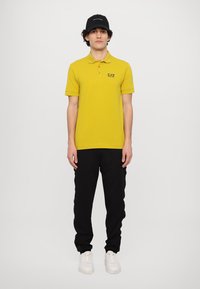Geel polo-shirt met korte mouwen en een zwart EA7-logo, gecombineerd met zwarte broek en witte sneakers, tegen een effen achtergrond.