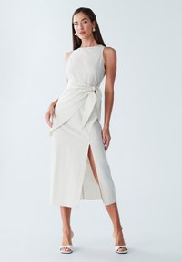 Midi-jurk in lichtbeige stof, met een boothals, detail met strik aan de zijkant en een hoge split tot op de dij. Soepele textuur en aansluitend silhouet.