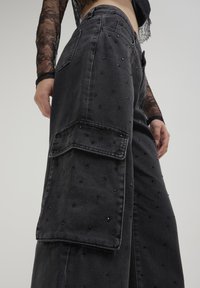 Pantaloni cargo in denim neri con design a gamba larga, caratterizzati da accenti testurizzati e perline decorative sul tessuto. Dettaglio della tasca laterale visibile.
