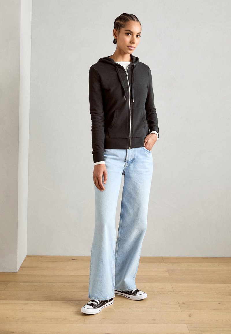 Black zip-up hoodie with front pockets, paired with light blue flared jeans. Model wears black and white sneakers. Simple, functional design.