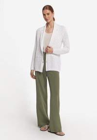 Weißer Strickcardigan mit Schalkragen, kombiniert mit einem beigen Oberteil und olivgrünen weiten Hosen. Verfügt über frontale Taschen und einen Knopfverschluss.