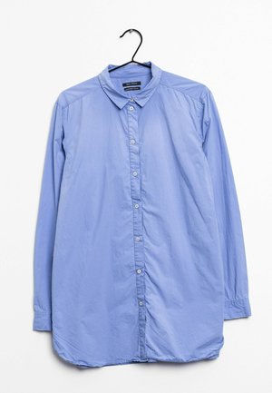 Chemise bleu clair à manches longues avec col et boutons, suspendue à un cintre noir sur un fond blanc.