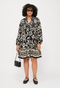 Robe à motifs noir et crème avec des manches longues, un ourlet froncé et un col. Accompagnée d'un petit sac à main noir et de chaussures plates.