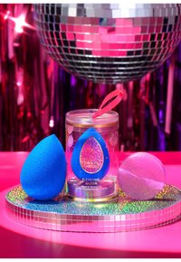 Blauwe make-up spons in een doorzichtige container met een roze lint. Inbegrepen is een ronde, iriserende reinigingszeep op een spiegelschaal.