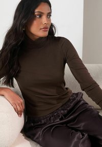 Brown ribbed turtleneck top paired with dark satin joggers, featuring a relaxed fit and drawstring waist, worn against a textured light background.