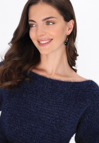 Granatowy, teksturowany sweter z dekoltem typu off-shoulder, w połączeniu z okrągłymi czarnymi kolczykami, na białym tle.