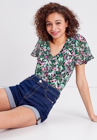 Blusa a maniche corte con sfondo nero e vivace motivo floreale in verde e rosa; abbinata a shorts in denim scuro con orlo arrotolato.
