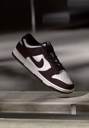 Scarpa da ginnastica Nike bianca e nera sospesa sull'aria sopra una panchina in cemento con ombre in un ambiente interno minimale.