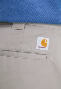 Beige bukser med et struktureret stof, der har et hvidt firkantet mærke med et gult og rødt logo på baglommen, paired med en blå fløjls top.