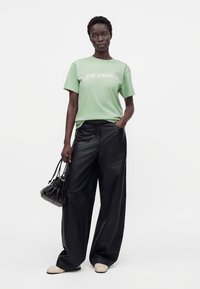 Personne portant un t-shirt vert pastel avec du texte, un pantalon en cuir noir ample, des chaussures beiges et portant un petit sac à main noir.
