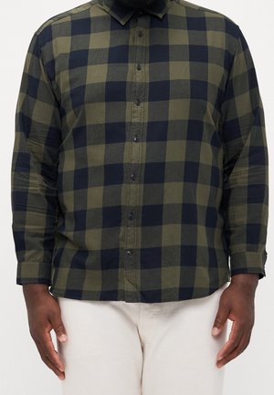 Uomo che indossa una camicia a quadri nera e verde oliva a maniche lunghe abbottonata con pantaloni beige, in piedi davanti a uno sfondo semplice.