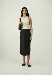 Top sem mangas em bege claro combinado com uma saia midi preta, segurando uma pequena mala preta. A modelo usa sapatos pretos planos e tem o cabelo liso.