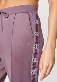 Μωβ αθλητική φόρμα με ριμπ ζώνη μέσης, πλαϊνές τσέπες και αντίθετη σατέν λωρίδα με μοτίβο λογότυπου κατά μήκος της εξωτερικής ραφής.