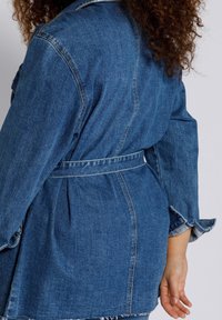 Jeansjacke in Mittelblau, tailliert mit einem Gürtel, mit lockerer Passform, umgeschlagenen Ärmeln und einem leicht ausgefransten Saum.