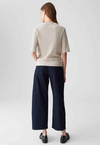 Haut en tricot beige à manches courtes et texture à motifs, associé à un jean large bleu marine. Des chaussures noires à enfiler complètent la tenue.