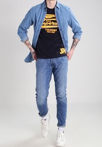 Mann trägt ein blaues Denimhemd über einem schwarzen Grafik-T-Shirt, blaue Jeans, weiße Sneaker und zeigt ein lässiges Full-Body-Outfit mit tätowierten Unterarmen.