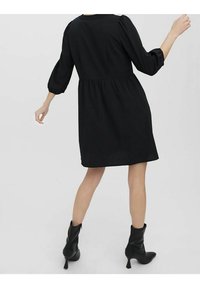 Robe noire longueur genou avec manches bouffantes, taille cintrée et jupe plissée. Portée avec des bottines à talon bloc. Texture de tissu lisse.