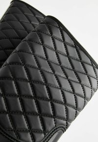 Sac en simili cuir matelassé noir avec un motif en diamant, des coutures visibles et une texture lisse. Dispose d'arêtes arrondies et d'un design élégant.