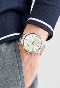 Silver and gold-tone watch with a white dial, three sub-dials, and a stainless steel band. Worn on a hand resting in a gray pocket.