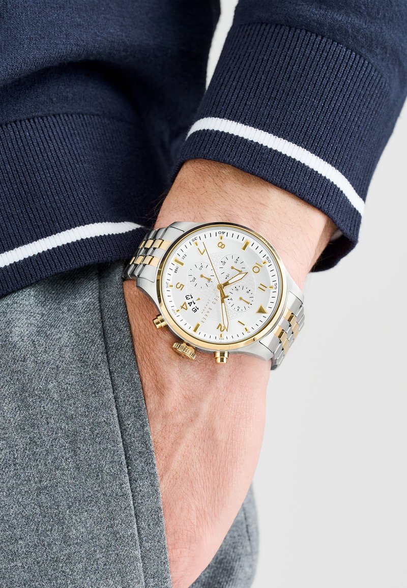 Silver and gold-tone watch with a white dial, three sub-dials, and a stainless steel band. Worn on a hand resting in a gray pocket.
