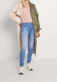 Abrigo trench color caqui con detalles beige, camiseta sin mangas rosa claro, jeans ajustados azules y zapatillas beige. Se pueden ver los botones del abrigo.