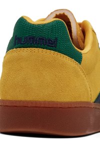 Žute suede tenisice s zelenim teksturiranim akcentima i smešnim gumenim đonom. Na peti je izvezen "hummel" logo.