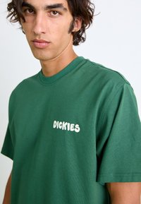 Grünes Baumwoll-T-Shirt mit Rundhalsausschnitt, das ein weißes "DICKIES"-Logo auf der Brust und kurze Ärmel aufweist. Glatte Textur, lässiges Design.