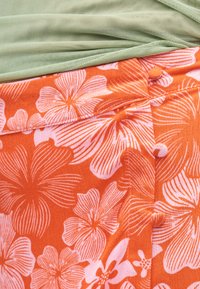Κοντινό πλάνο πορτοκαλί υφάσματος με μεγάλo ροζ λουλουδάτο σχέδιο και κουμπιά καλυμμένα με ύφασμα, σε συνδυασμό με ανοιχτοπράσινο υφαντό ύφασμα.