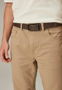 Beige katoenen broek met een slim fit en subtiele textuur, gecombineerd met een bruine leren riem met een rechthoekige metalen gesp.
