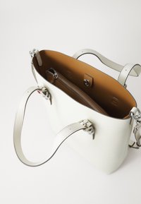 Bijela kožna torba s aluminijskim detaljima, sadrži zip džep unutra i kontrastnu smeđu podstavu. Dvostruke ručki za nošenje.