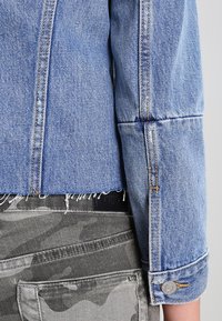 Veste en denim bleu clair avec un design court, un ourlet effiloché et des poignets retroussés. Associée à un pantalon en camouflage gris avec des poches standard.