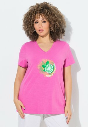 Vrouw met krullend haar draagt een felroze T-shirt met V-hals met een schijfje citrusvrucht en de tekst "Slice of Sunshine", gecombineerd met witte broek.