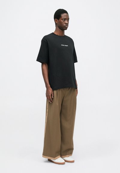 Schwarzes T-Shirt mit kurzen Ärmeln und weißen gestickten Text, kombiniert mit weiten beigen Hosen und weißen Sneakers mit braunen Sohlen.